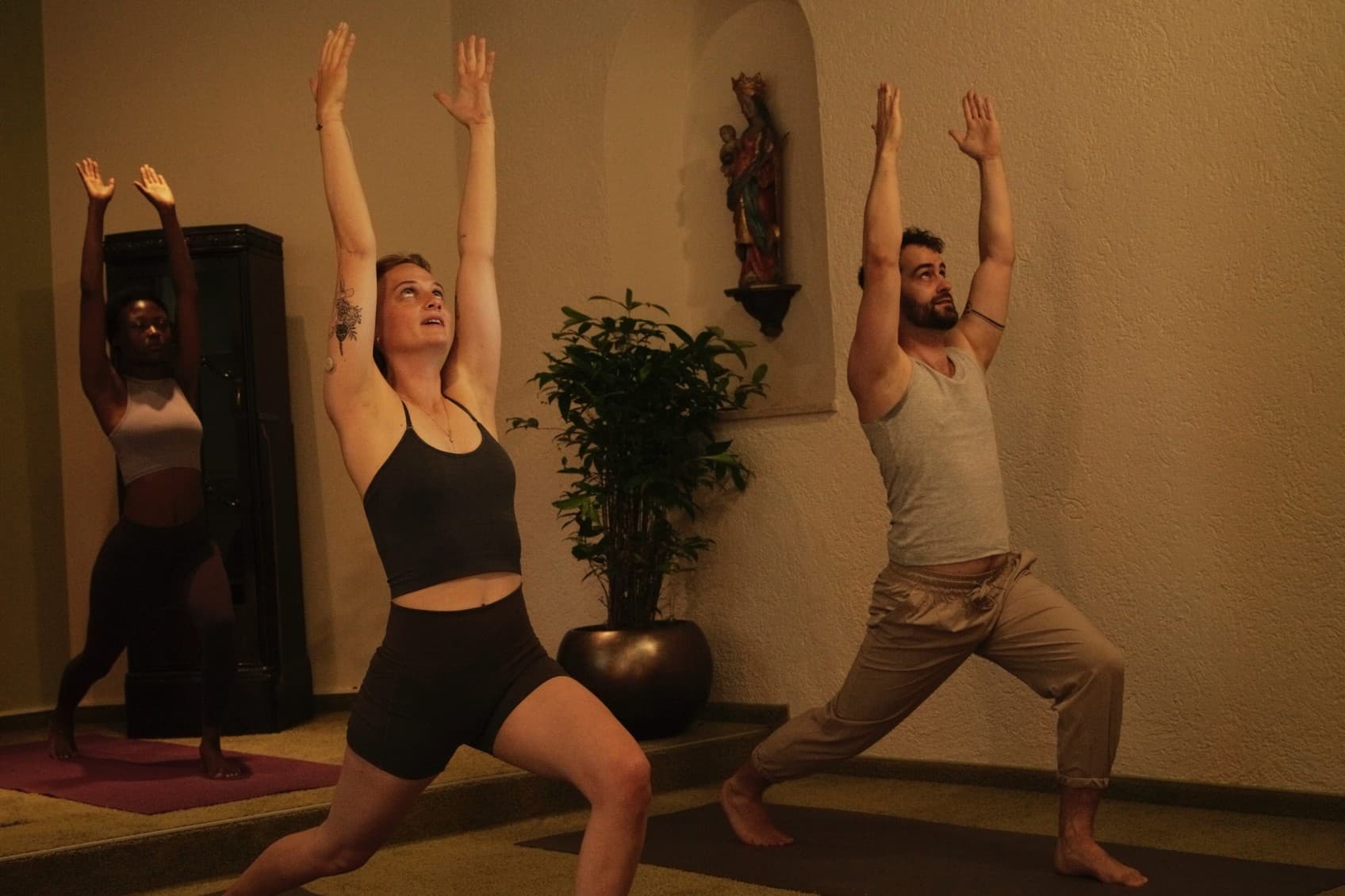 Zu sehen sind zwei Personen, die an einer Yoga-Stunde teilnehmen. Sie nehmen eine Haltung ein, bei der sie die Arme heben und ausatmen. Die Szene spielt in einem Yoga-Raum im Hideauts Hotel „Der Rosengarten“.