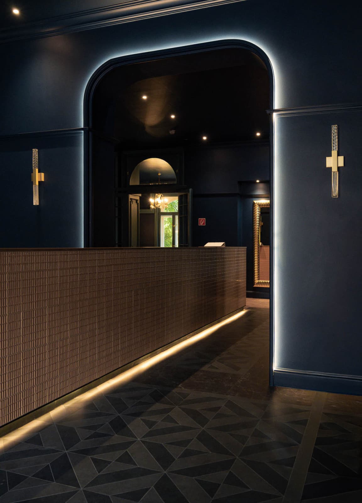 Seen is lobby, which is seperated in two by a lit up archway, that compliments the dark blue design of the hotel. In the middle is a long small wall, also seperating the lobby in two parts.