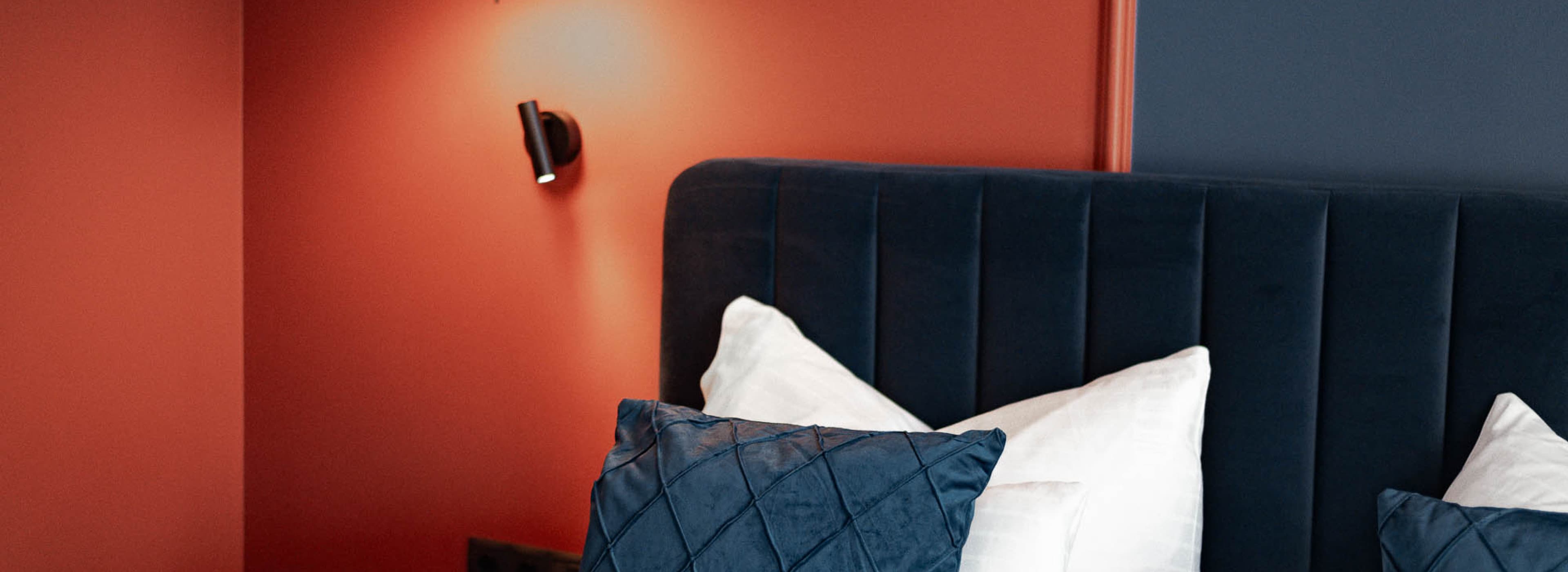 Seen is a sample room detail picture of a room, which shows the bed and bedframe. The colours orange and blue are seen in the background on the walls.