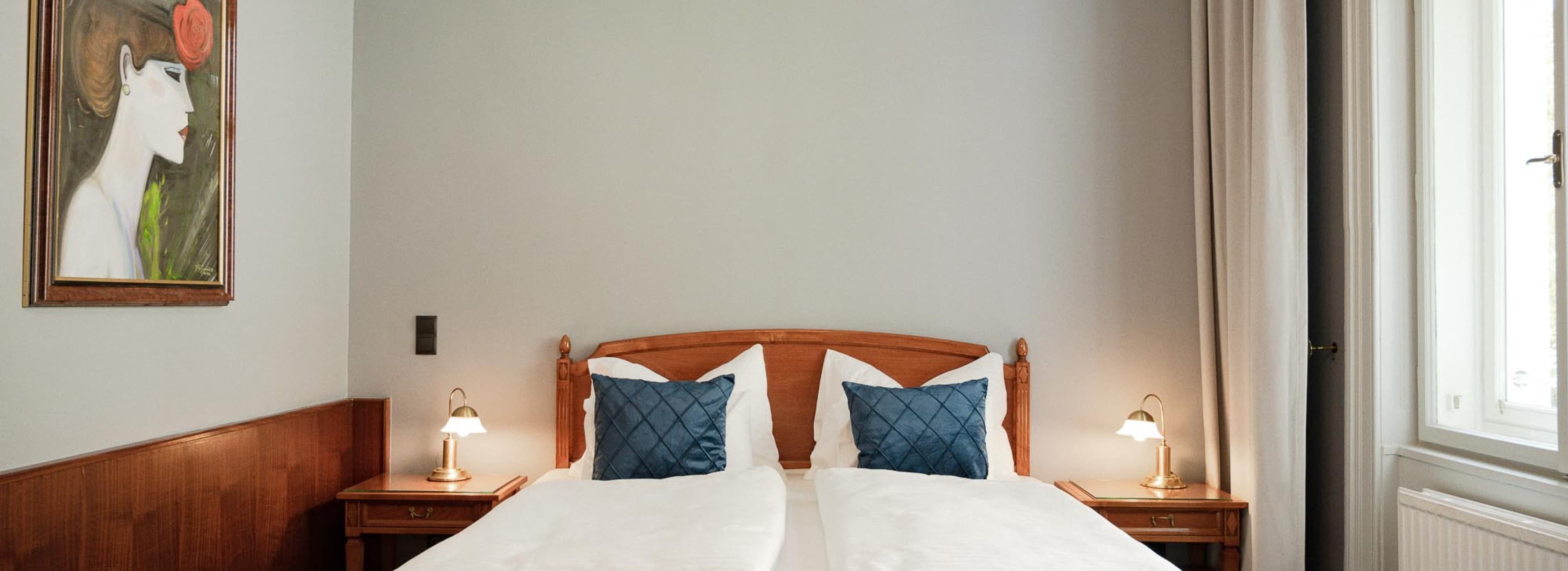 The picture is showing the double bed side of the room. The category comfy is for two people and offers a small couch as well. On the left side a painting of a woman in a art deco style is seen.