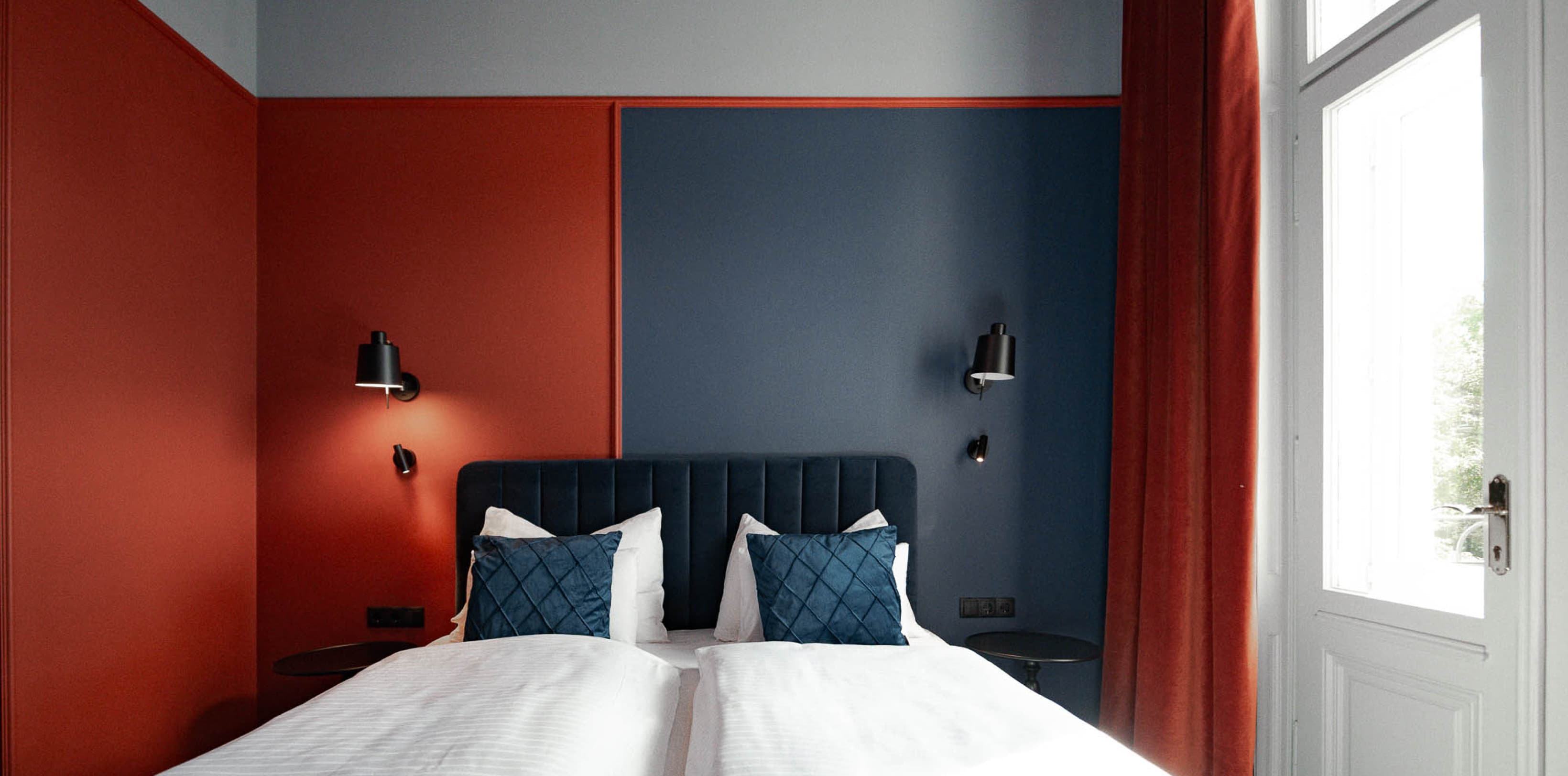 Auf diesem Bild ist das Doppelbett zu sehen. Die Hälfte des Raumes ist dunkelblau gestrichen, die andere Hälfte orange. Auf der rechten Seite ist auch das restaurierte Fenster zu sehen.